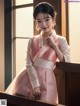 A woman in a pink and white hanbok standing by a window.