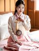 A woman in a pink and white hanbok sitting on a bed.