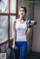 A woman in a white tank top and blue leggings holding two blue dumbbells.